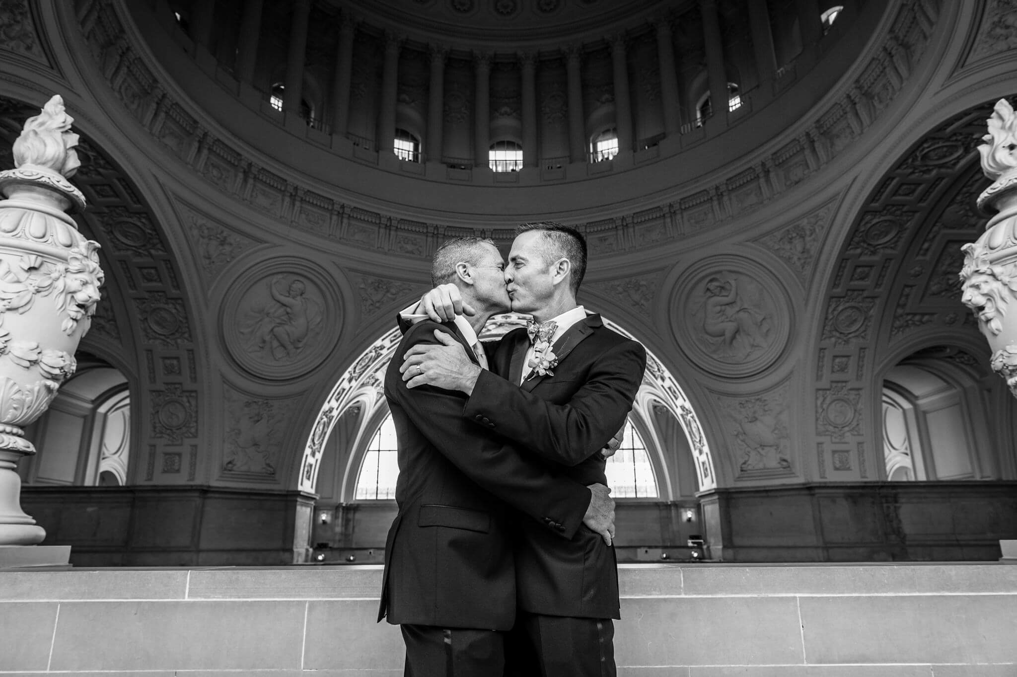 A black and white photo of two grooms kissing under architectural dome at the San Francisco City Hall