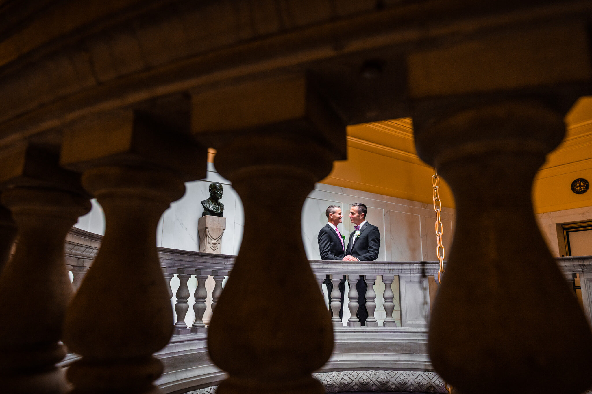 Color photo taken through balusters of two men holding hands and embracing on balcony of San Francisco City Hall Wedding