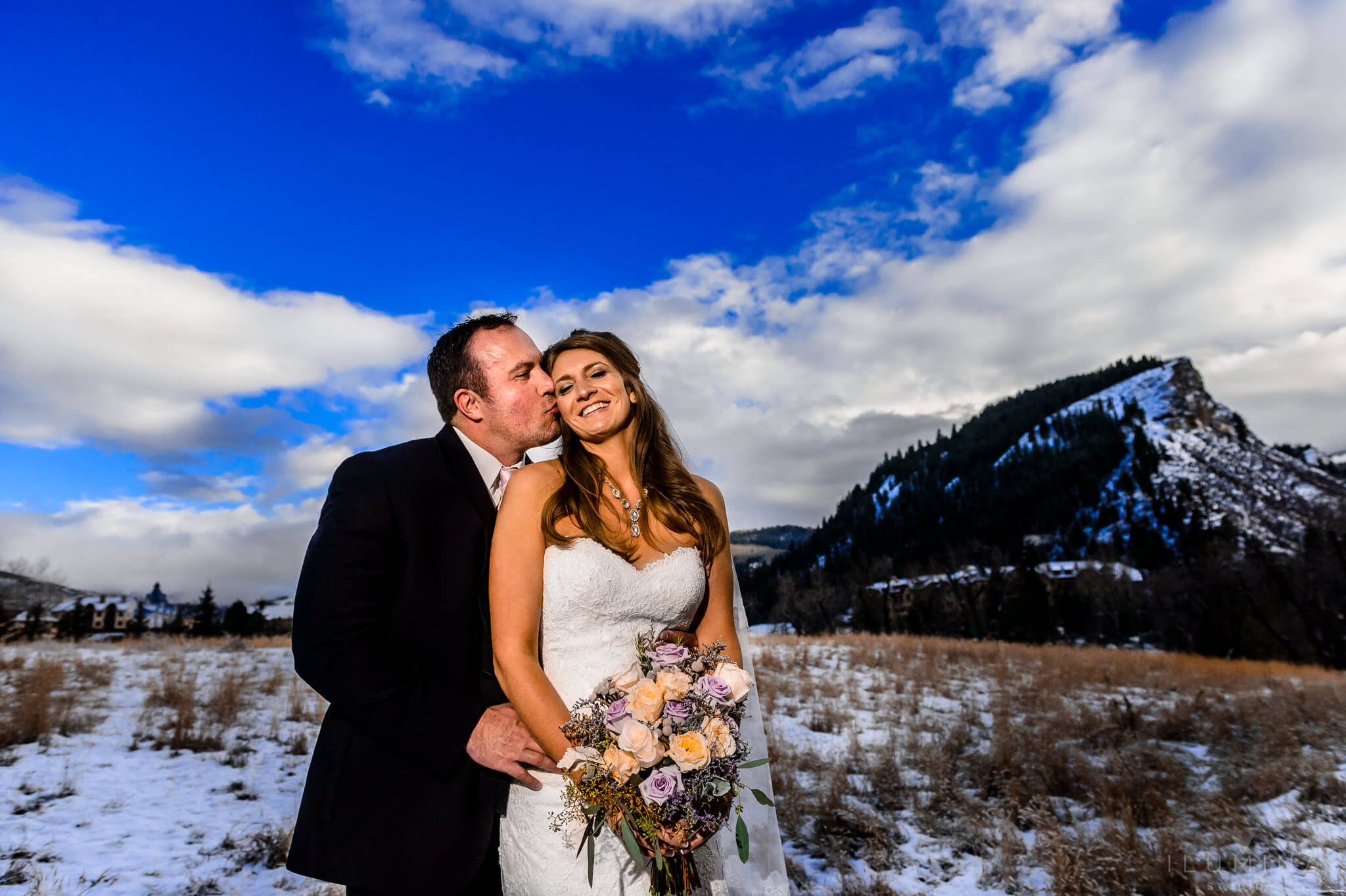 Color photo of groom kissing bride's cheek with snowy Vail landscape in background