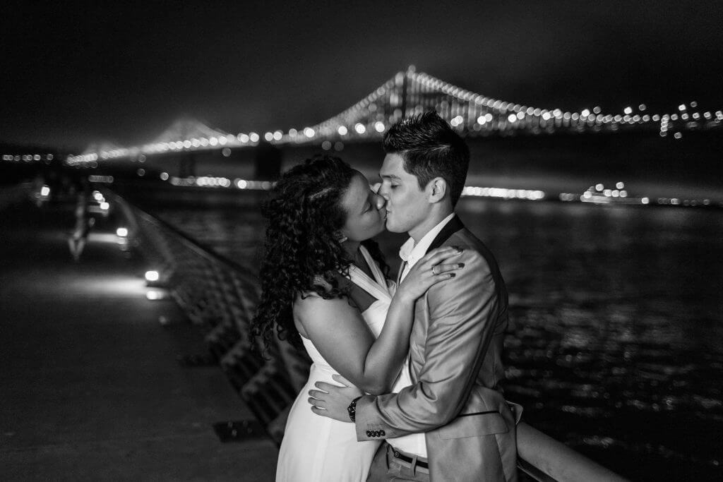 Black and white documentary wedding photo of bride kissing groom in front of Golden Gate Bride at nighttime in San Francisco