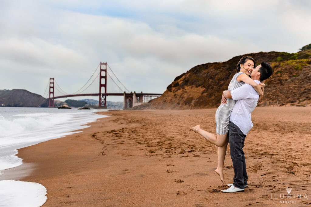 Color engagement photo of man hug-lifting barefoot woman on beach next to Golden Gate Bridge in San Francisco, California