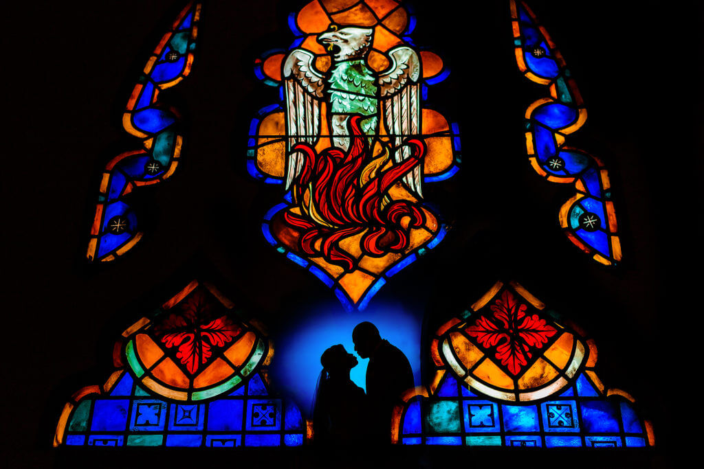 Color wedding photograph of stained glass with silhouette of bride and groom leaning in to kiss