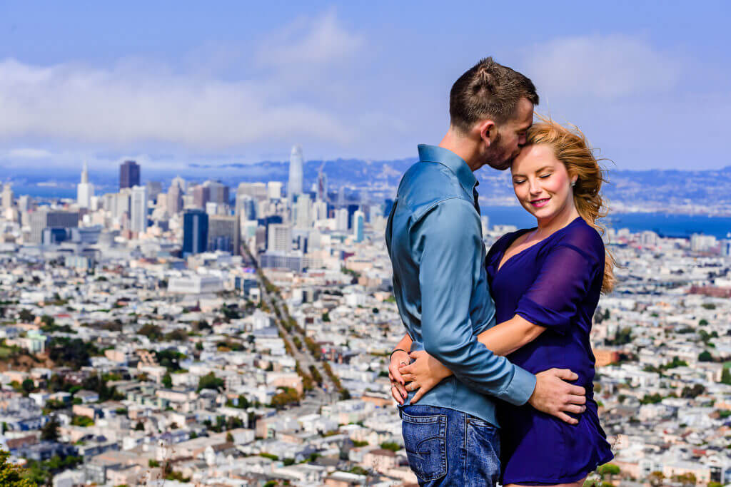 Engagement Photos in San Francisco. Color photograph of man kissing woman's temple as they hug each other in front of San Francisco cityscape background during engagement photoshoot