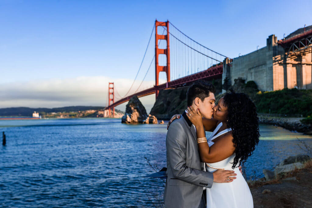 Color photograph of bride and groom kissing by the San Francisco Bay with Golden Gate Bridge in background during San Francisco, California wedding shoot