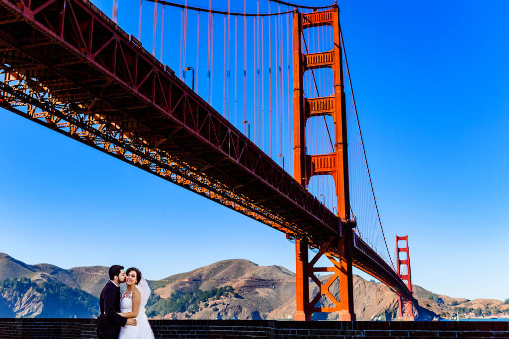 Color wedding photo of groom kissing bride's cheek below the Golden Gate Bridge in San Francisco, California