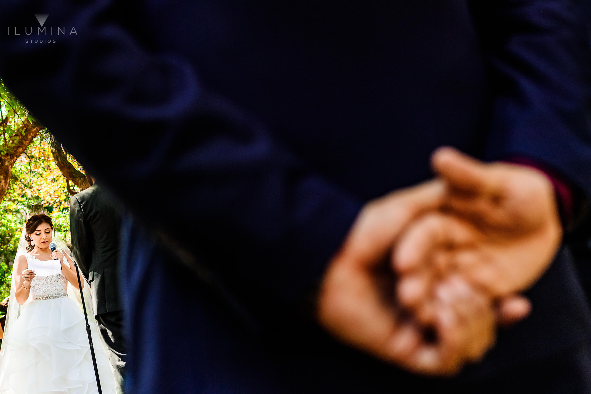 Color documentary-style image of bride reading vows to groom in background with groomsman's hands in foreground at Anaheim Oak Canyon Nature Center wedding