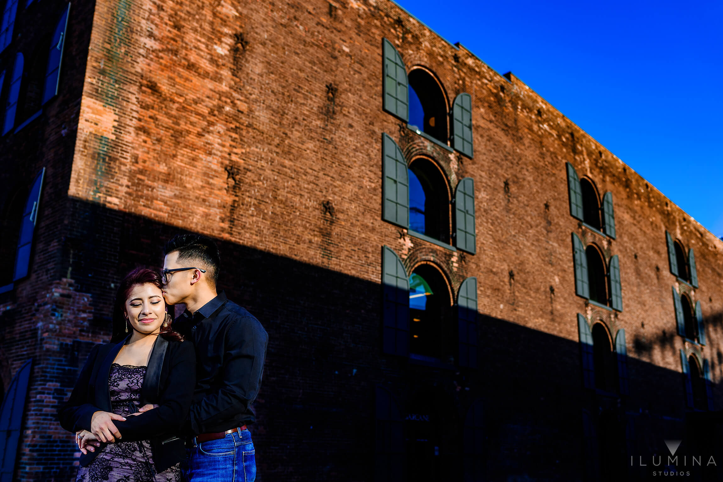Color photo of man kissing fiancée's temple in front of large brick building in sunny Brooklyn, New York City
