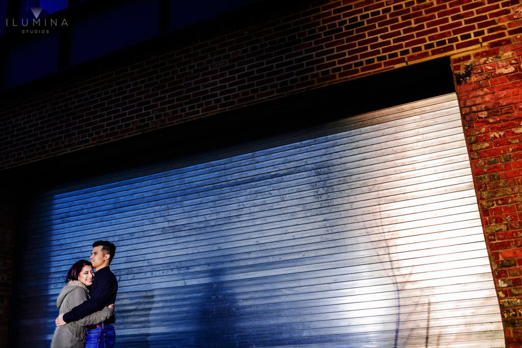 Man and woman smile and hug in front of brick building and metal garage door in Brooklyn, New York