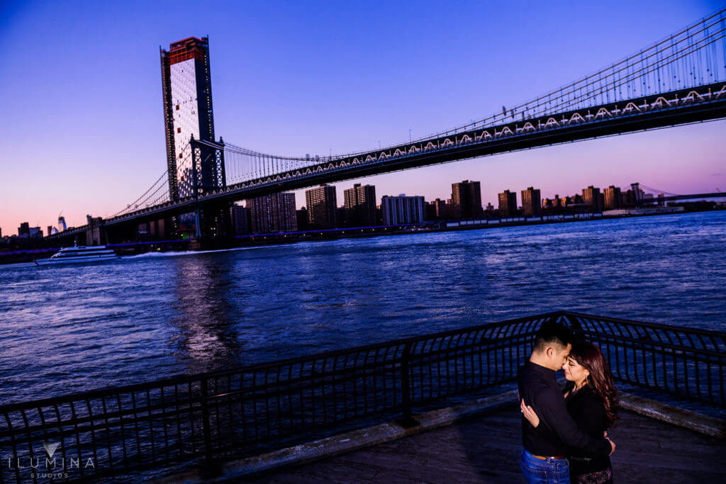 Man and woman hold each other in front of the East River and Manhattan Bridge in Brooklyn, New York