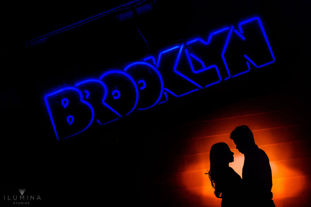 Multiple exposure engagement portrait of male and female silhouettes embracing inside red spotlight with blue Brooklyn sign above in Brooklyn, New York