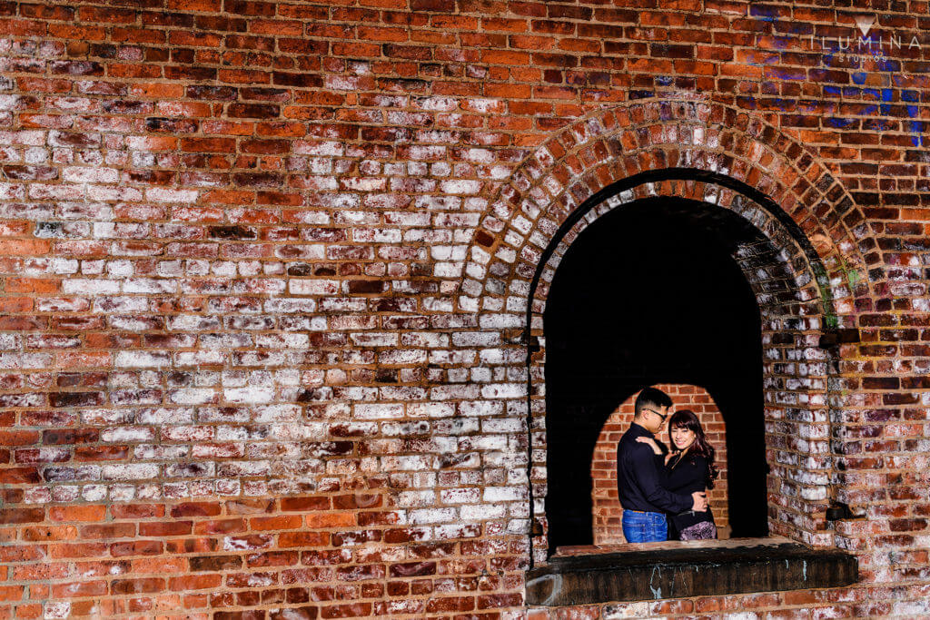 Engagement photo of man and woman embracing behind open brick window in Brooklyn, New York City