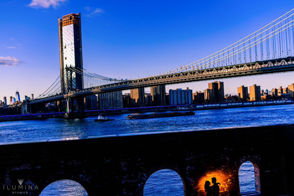 Color photo of Manhattan Bridge with small silhouette of man and woman embracing in foreground