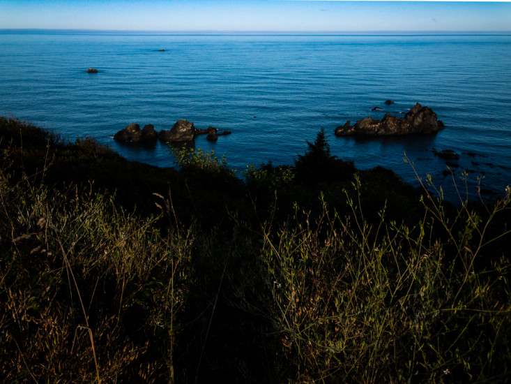 From high on a grassy hill looking down to still blue waters with scattered large rocks below