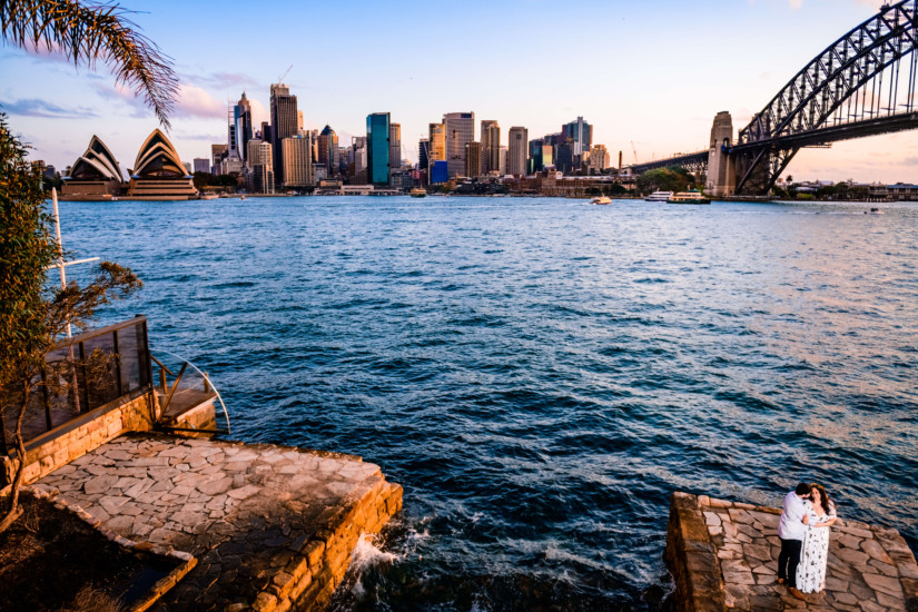 Man and woman standing on a stone pathway overlooking the wave crashing water as the city and sydney opera house rest in the background