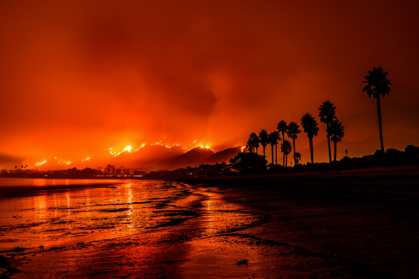 Large fire in the sky near the beach, everything a shade of red with silhouettes of palm trees on the right side