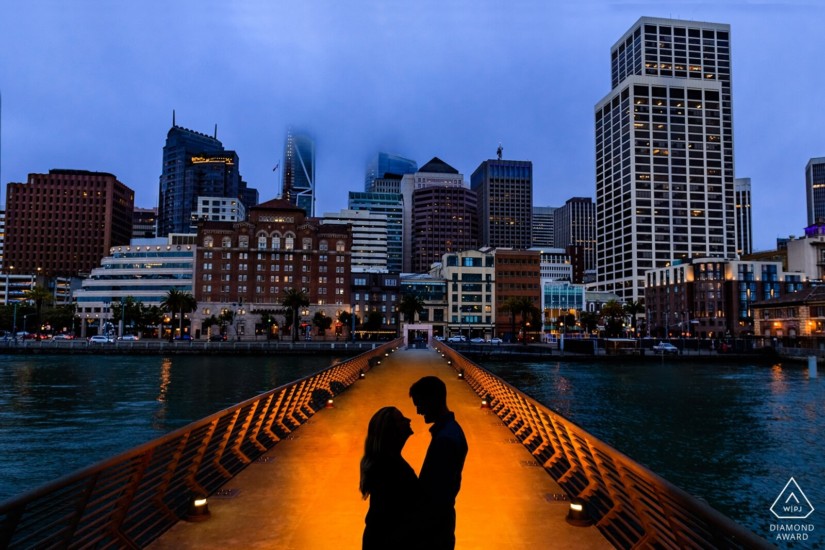 man and woman silhouette embrace on a long pier near the city, over a river