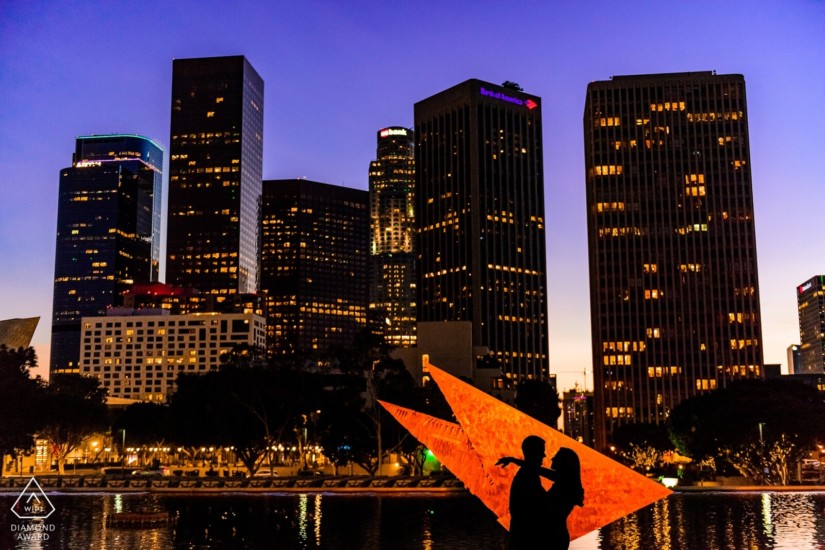 man and woman silhouette she has her arms wrapped around him and gaxing into his eyes in front of a city with towering buildings overlooking the river