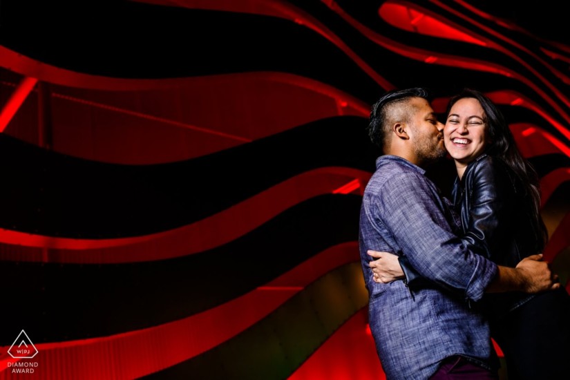 man and woman holding eachother he is kissing her cheek and she has a large happy smile on her face. black and red wavy background behind them